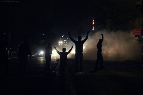 Protestors hold up peace signs as the headlights from two water cannon trucks shine on them.